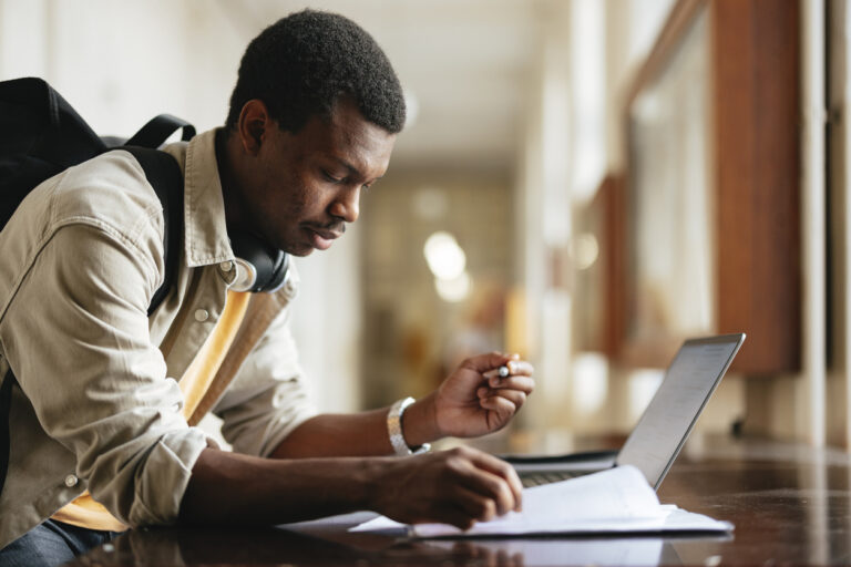 University student wearing backpack consults laptop while filling out paperwork.
