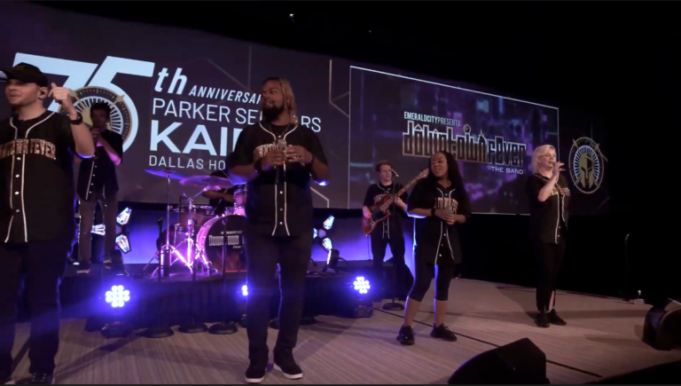 Several singers and musicians in baseball jerseys perform in front of a screen that reads "75th Anniversary of Parker Seminars--Kairos"
